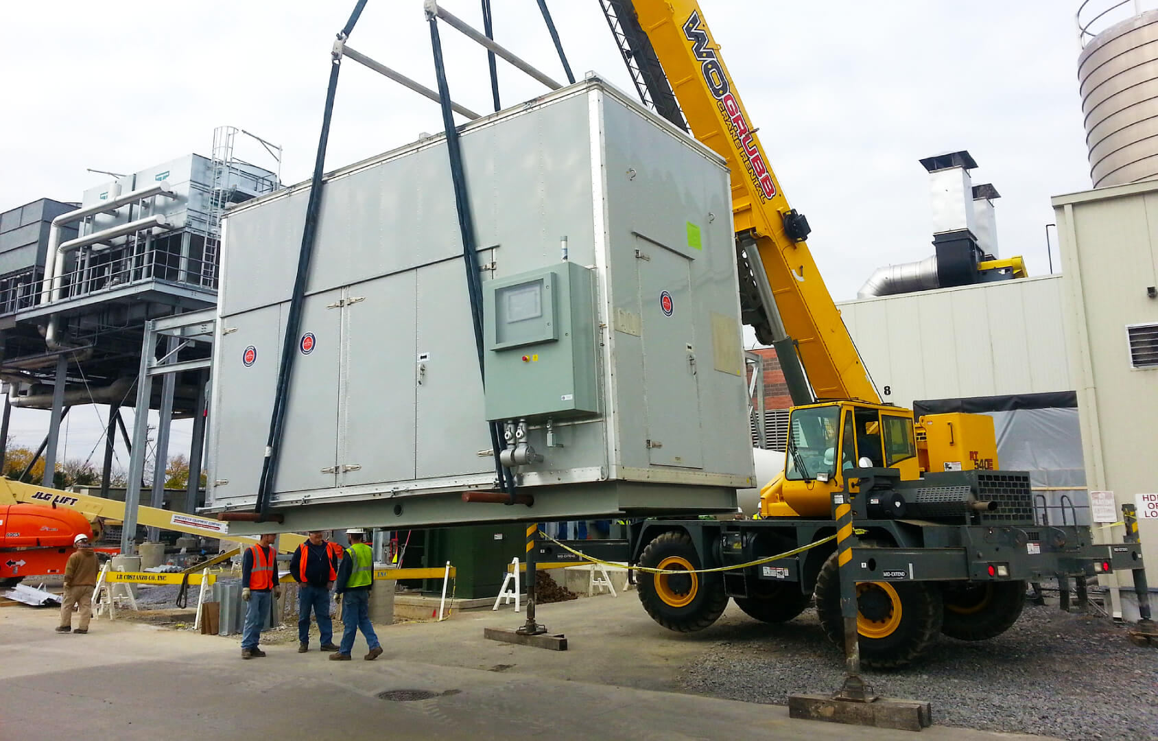 A yellow crane lifts a large industrial unit while several workers in safety vests and helmets stand nearby. The scene takes place at an industrial facility with machinery and buildings in the background.