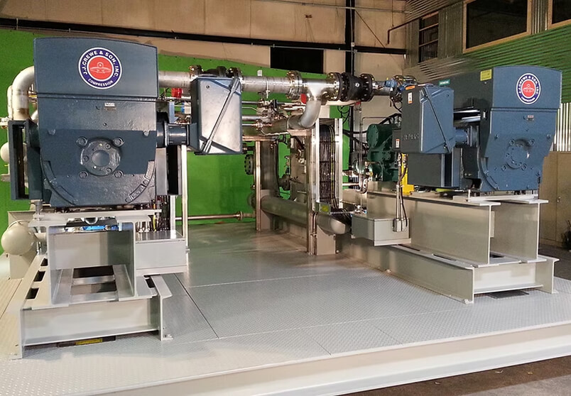 Industrial equipment featuring two large blue compressor units with labels, connected by metal pipes and mounted on a raised metal platform inside a factory with a green wall in the background.