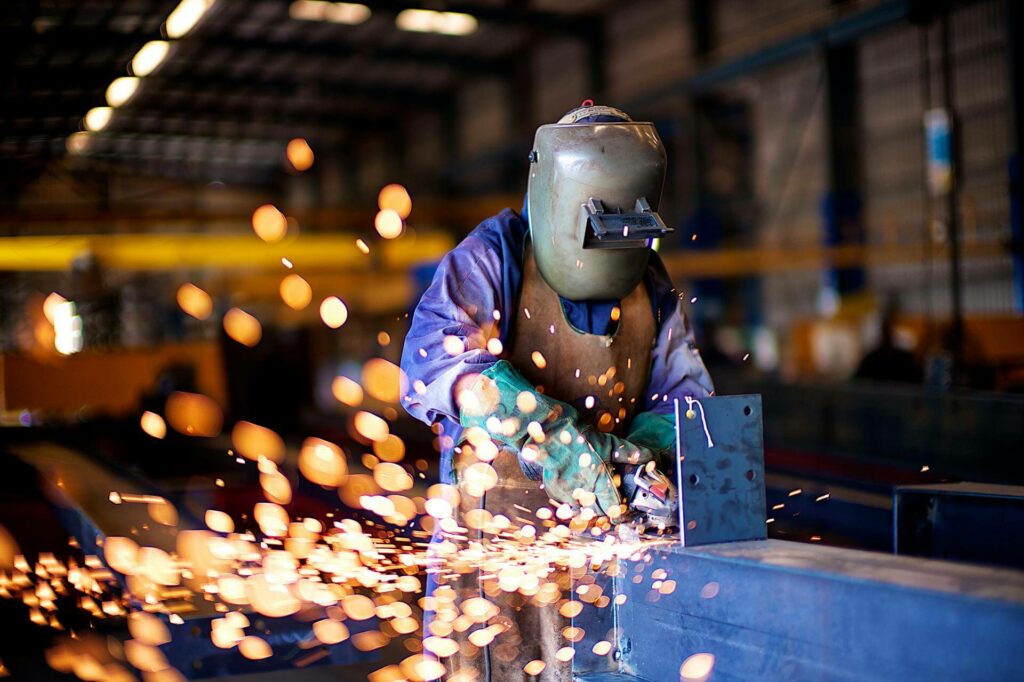 A worker wearing protective gear, including a welding helmet and gloves, uses a tool to grind or cut metal, creating bright, flying sparks in an industrial workshop setting.