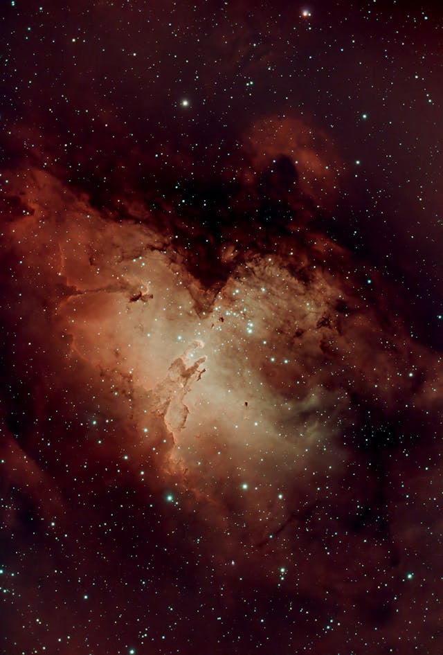 A nebula in deep space glows with red, orange, and cream clouds of gas and dust, surrounded by numerous bright stars scattered across a dark sky.