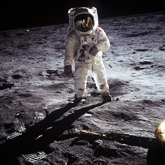 An astronaut in a white suit stands on the rocky, dusty surface of the Moon; footprints are visible on the ground, with dark shadows stretching behind the astronaut and black space in the background.